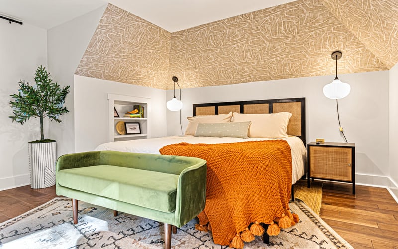 Bedroom with green velvet bench, queen bed with orange throw, rattan nightstand, and sloped ceiling with geometric wood panels