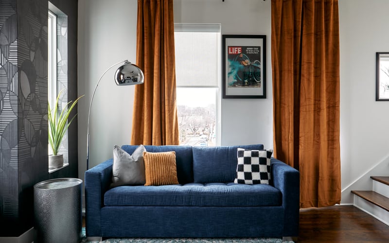 Blue sofa with patterned pillows facing tall windows with rust-colored curtains and framed artwork on white walls