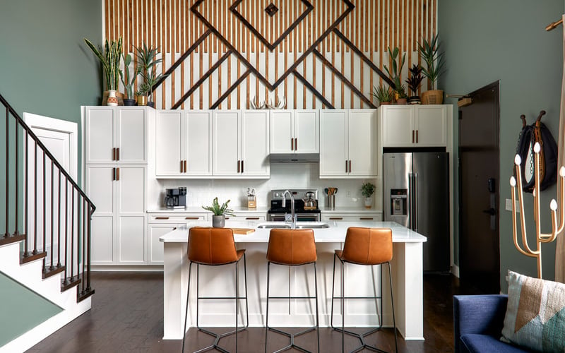Modern kitchen with white cabinetry, stainless steel appliances, and three orange leather bar stools at island, geometric wood wall art above