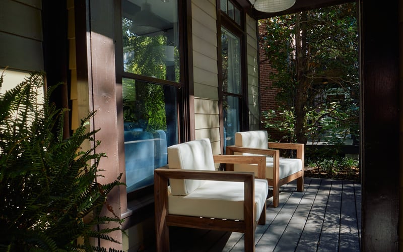 Covered patio with wooden chairs, white cushions, and surrounding greenery and climbing vines