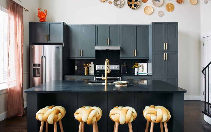 Modern kitchen with dark gray cabinetry, gold hardware, stainless steel appliances, black island with four woven wood stools, and decorative plates on white