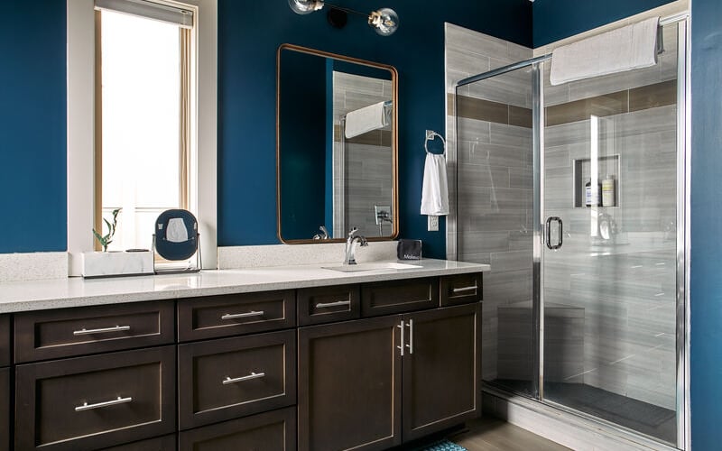 Modern bathroom with deep blue walls, dark wood vanity, white countertop, framed mirror, and glass-enclosed shower with gray tile.