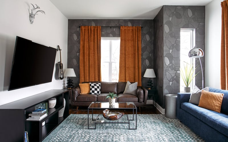 Modern living room with dark gray patterned walls, brown leather sofa, orange curtains, flat-screen TV, and blue armchair