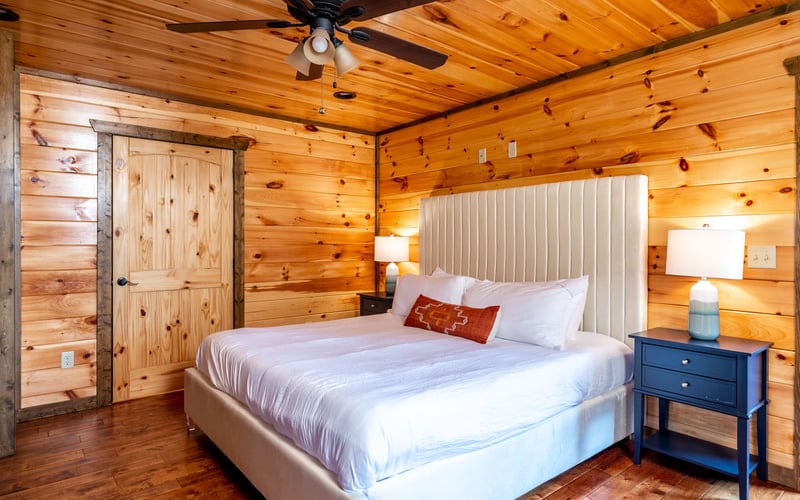 Wooden cabin bedroom with queen bed, white headboard, blue nightstand, and ceiling fan