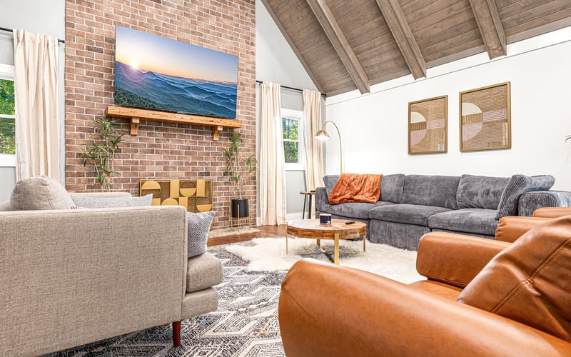 Modern living room with exposed brick fireplace, gray sectional sofa, tan armchair, and mountain landscape artwork
