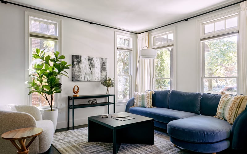 Bright living room with navy sectional sofa, black coffee table, cream armchair, and multiple windows overlooking trees