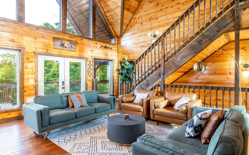 Spacious wooden cabin living room with blue and tan seating, round black table, and metal staircase to loft