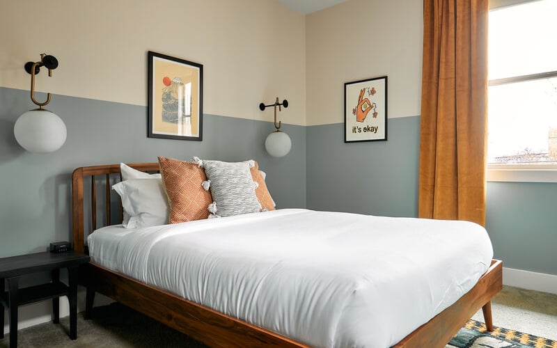 Bedroom with wooden bed, white linens, two-tone blue and cream walls, framed artwork, and orange curtains