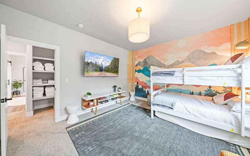 Bright bedroom with white bunk beds, mountain landscape mural, framed nature photo, low shelf, and open shelving unit with towels