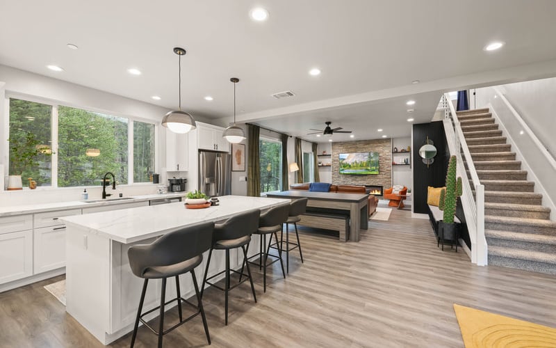 Open-plan kitchen and living area with white cabinetry, island seating, staircase, and large windows overlooking greenery